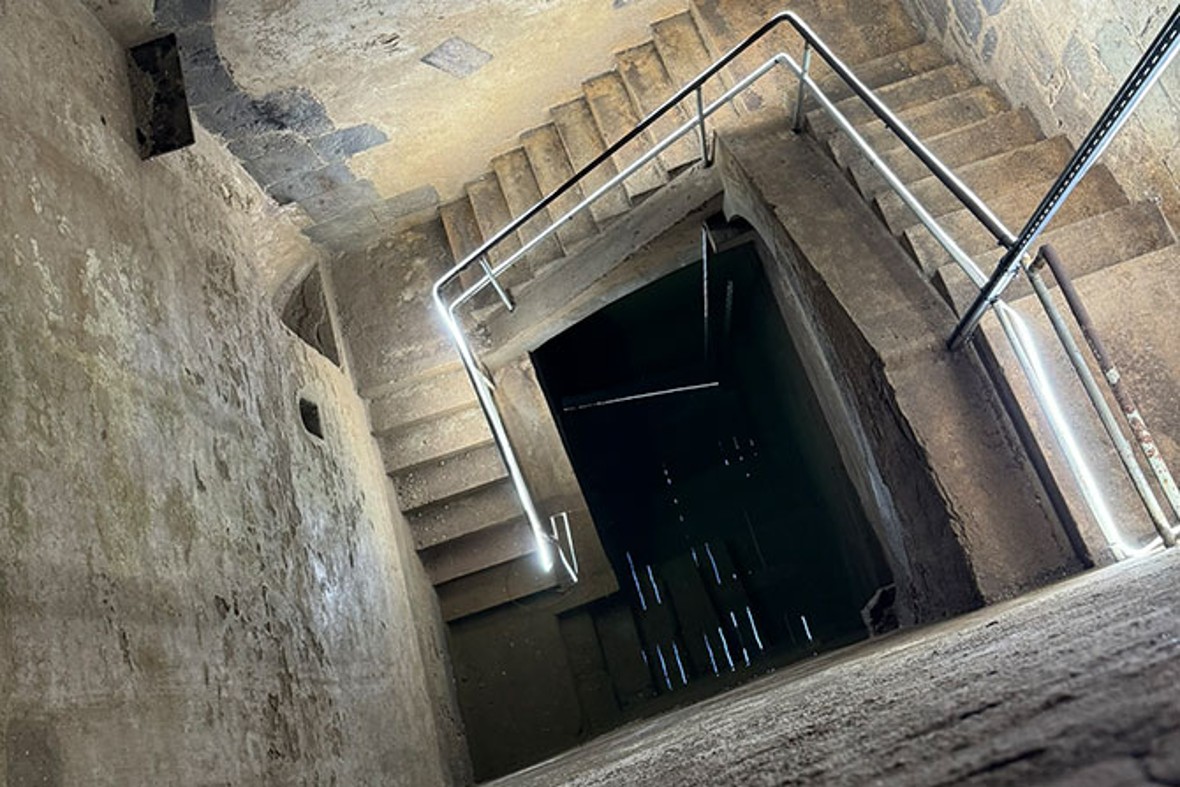 Die Aufnahme zeigt einen steinernen Treppenabgang in einem historischen Bauwerk, vermutlich eine Mikwe oder ein unterirdisches Ritualbad. Die Treppen sind steil und verlaufen in mehreren Abschnitten spiralförmig nach unten. Rostige Metallgeländer entlang der Stufen deuten auf das Alter der Konstruktion hin. Die Wände bestehen aus grob behauenen Steinen und teilweise verputzten Flächen, die von der Zeit gezeichnet sind. Der Boden ist aus massiven Steinplatten gefertigt, und die Beleuchtung bringt die Textur und Patina der Wände sowie die Spuren der Vergangenheit eindrucksvoll zur Geltung. Die Tiefe des Schachts vermittelt ein Gefühl von Enge und archäologischer Bedeutung.