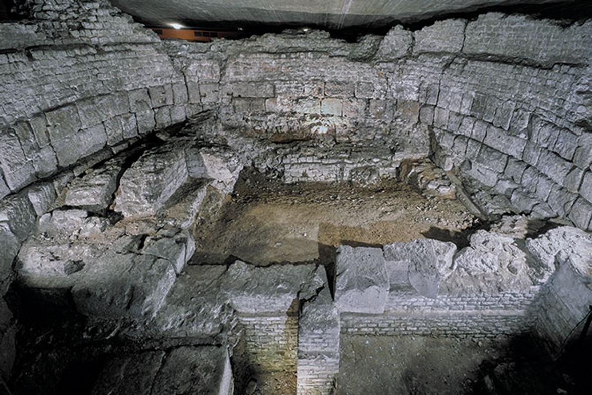 Die Aufnahme zeigt die Überreste einer archäologischen Ausgrabungsstätte, vermutlich römischen Ursprungs. Massive Steinblöcke formen die Wände einer antiken Struktur, deren Mauerwerk teilweise gut erhalten ist. Der Raum ist halbrund angelegt, ähnlich einem Amphitheater oder einer Therme, und die Spuren von Fundamenten und Wandsegmenten sind deutlich sichtbar. Der Bodenbereich ist freigelegt, mit Erdspuren und verwitterten Steinfragmenten. Die Atmosphäre wirkt gedämpft, vermutlich durch künstliche Beleuchtung während der Dokumentation der Fundstätte. Die Architektur und die Größe der Steine deuten auf eine bedeutende antike Konstruktion hin.